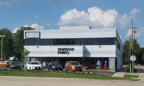 A two-story building with a sign reading "Haberman Fabrics," featuring parked cars and a blue sky with clouds.
