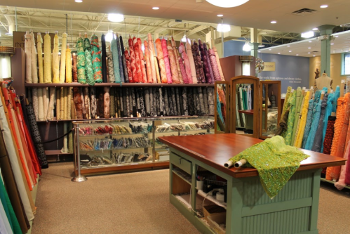 A fabric store interior featuring colorful rolls of fabric stacked on shelves and a central cutting table.