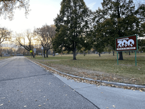 A road leading to Dakota Zoo, surrounded by trees and fallen leaves, with a sign indicating the zoo's entrance.