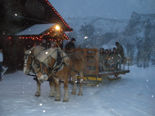 A horse-drawn sleigh with passengers in a snowy landscape, near a lit cabin and mountains in the background.