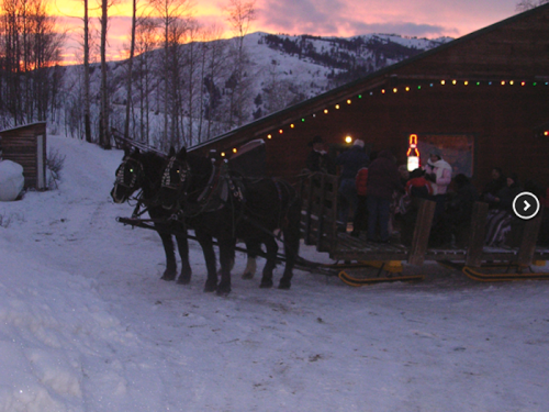Two horses stand in the snow near a wooden building, with people gathered inside and festive lights glowing at sunset.