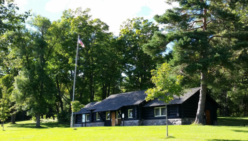 A log cabin surrounded by trees, with a flagpole displaying the American flag in a grassy area.