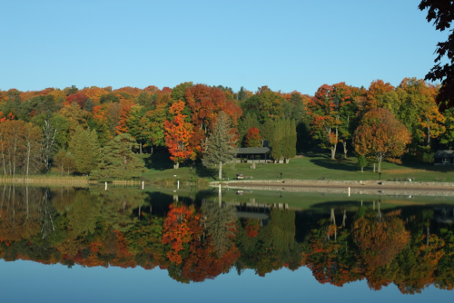 A serene lake reflects vibrant autumn foliage and a cozy cabin amidst colorful trees under a clear blue sky.