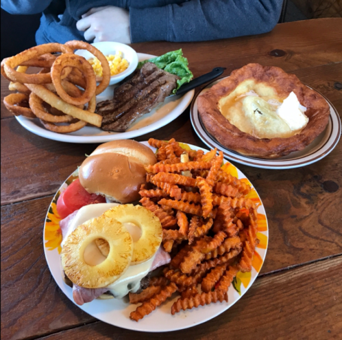 A table with three plates of food: curly fries, steak, and a dessert with a flaky crust and butter, plus burgers and sweet potato fries.