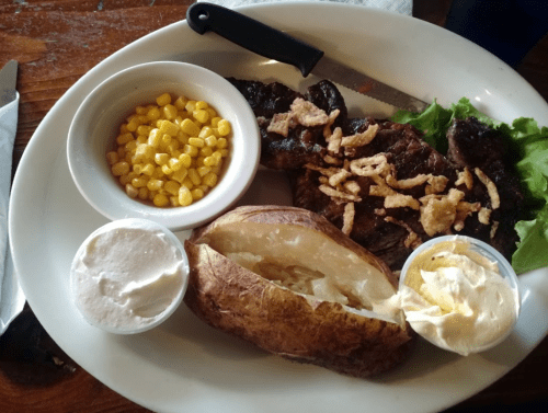 A plate with grilled steak, corn, a baked potato with butter, and crispy onions on lettuce.