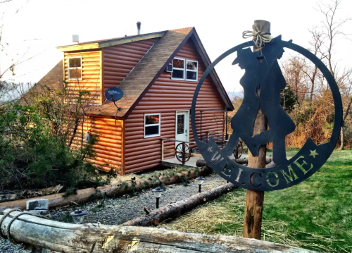 A rustic log cabin with a "Welcome" sign in front, surrounded by greenery and a clear sky.