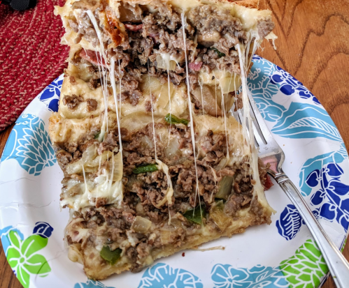 A slice of cheesy lasagna with layers of ground beef, vegetables, and melted cheese on a paper plate.