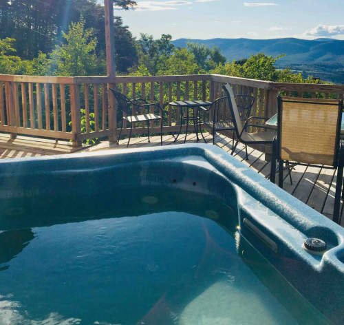 A hot tub on a deck overlooking a scenic mountain view with trees and blue sky in the background.