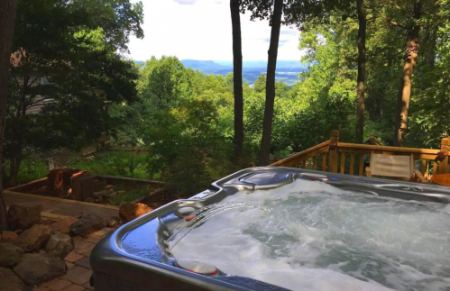 A hot tub on a wooden deck surrounded by trees, overlooking a scenic view of mountains and valleys.