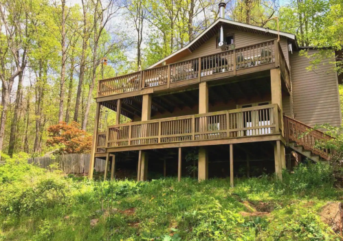 A wooden cabin with a large deck, surrounded by lush green trees in a serene forest setting.