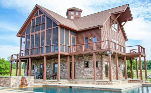 A large, modern wooden house with a stone base, featuring a screened porch and a pool in front.
