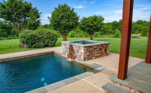 A serene backyard with a stone hot tub, swimming pool, and lush green landscaping under a clear blue sky.