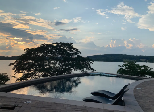 A serene lakeside view at sunset, featuring a pool and lounge chairs with trees silhouetted against the sky.