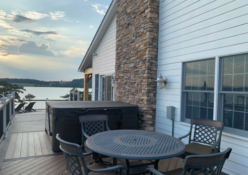 A lakeside deck with a hot tub, dining table, and chairs, surrounded by a scenic view of the water and sunset.