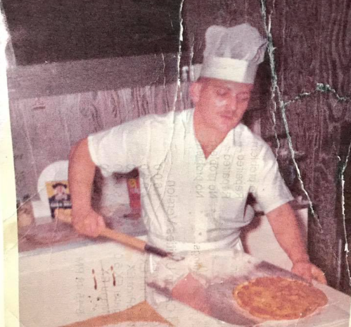 A chef in a white uniform and hat prepares a pizza in a kitchen, smiling as he works.