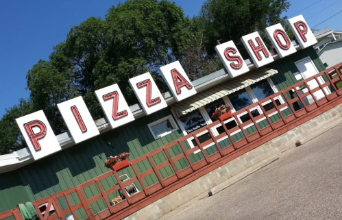 A pizza shop with a large, bright sign and a wooden fence, surrounded by greenery on a sunny day.