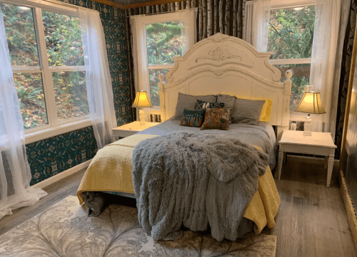 Cozy bedroom with a white bed, yellow accents, plush gray throw, and large windows overlooking greenery.