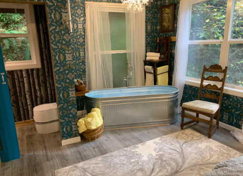 A stylish bathroom featuring a metal tub, patterned wallpaper, a chair, and large windows with natural light.