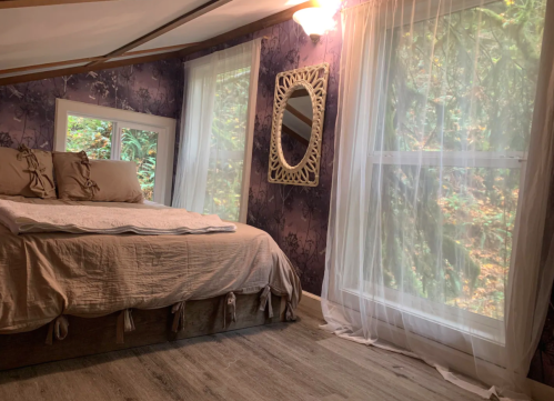 Cozy bedroom with a bed, decorative mirror, and large windows draped with sheer curtains, surrounded by greenery.