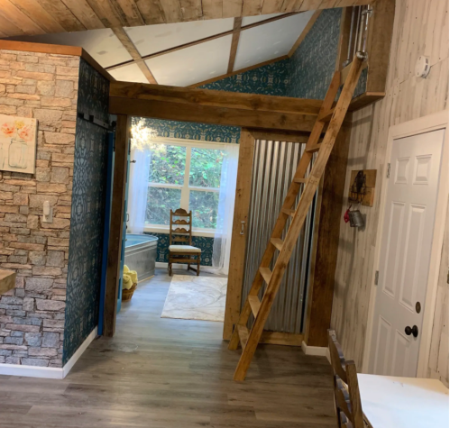 Interior view of a cozy room featuring wooden beams, a ladder, and a stone accent wall, with natural light from a window.