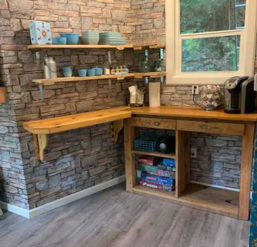 A cozy kitchen corner with wooden shelves, blue dishware, and a coffee maker against a stone-patterned wall.