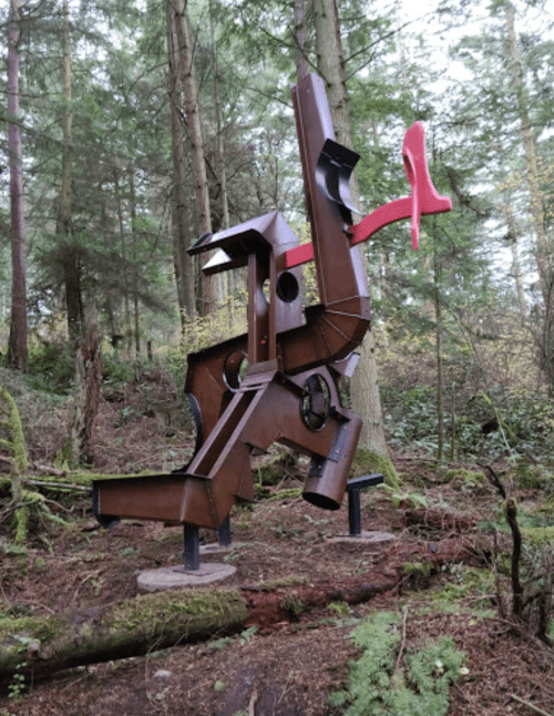 A large abstract metal sculpture stands among trees in a forest, featuring geometric shapes and a red accent.