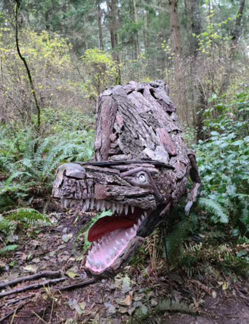 A whimsical wooden sculpture of a wolf's head, surrounded by lush greenery in a forest setting.