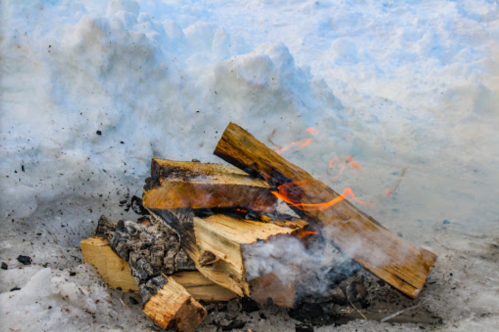 A small fire burns on a pile of logs, surrounded by snow and smoke rising into the air.