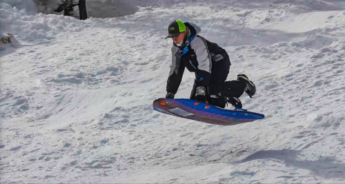 A child in winter gear jumps on a sled, soaring over a snowy hill.
