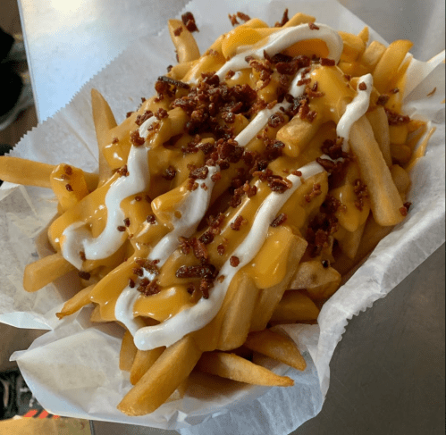 A serving of loaded fries topped with melted cheese, sour cream, and crispy bacon bits in a paper tray.