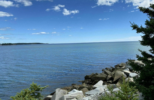 A serene coastal view featuring calm blue waters, rocky shoreline, and a clear sky with scattered clouds.