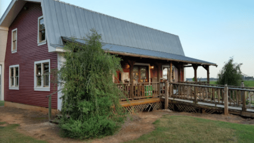 A red farmhouse with a metal roof, featuring a wooden deck and surrounded by greenery, set against a clear sky.