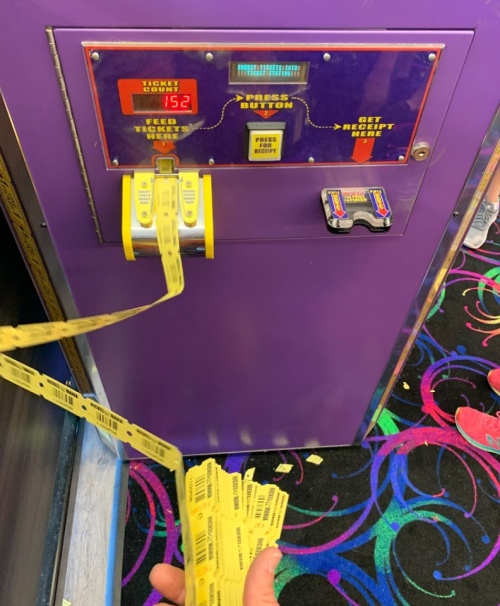 A ticket redemption machine dispensing long rolls of tickets, with a digital display showing "152" and colorful carpet below.
