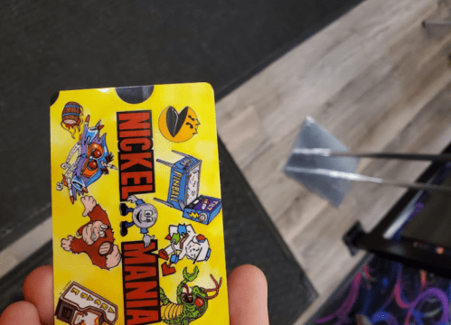 A hand holding a yellow arcade card featuring colorful cartoon graphics and the text "NICKEL MANIA."