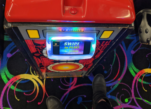 A colorful arcade game machine with a "Swipe to Play" screen, set against a vibrant, patterned carpet.