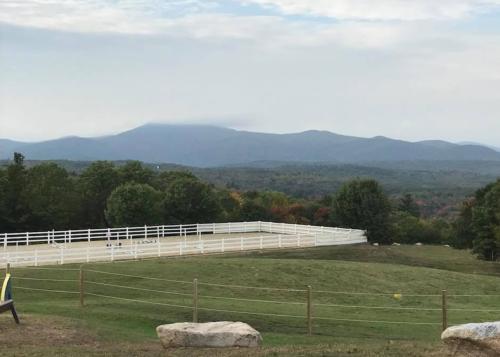 A scenic view of rolling hills and mountains under a cloudy sky, with a white fenced area in the foreground.