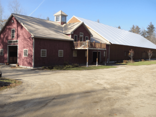 A large red barn with a metal roof, featuring a balcony and surrounded by trees and a gravel path.