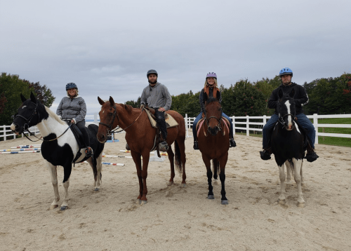 Four riders on horseback in an outdoor arena, wearing helmets and casual attire, with a cloudy sky in the background.