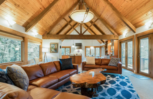 Cozy living room with wooden beams, leather sofas, a round coffee table, and large windows overlooking nature.