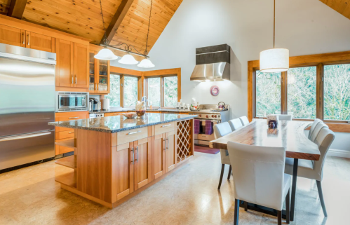 Modern kitchen with wooden cabinets, granite island, stainless steel appliances, and a dining table by large windows.