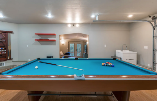 A bright game room featuring a blue pool table, cue stick, and a view of a cozy seating area in the background.
