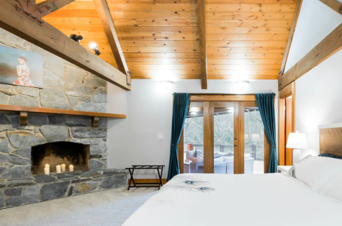 Cozy bedroom with wooden beams, stone fireplace, and large windows leading to an outdoor seating area.