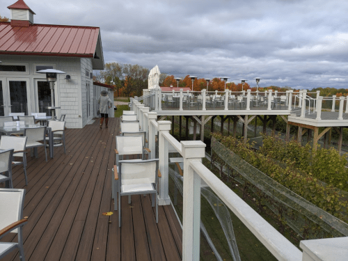 A wooden deck with white chairs overlooks a scenic view, with a person walking away and cloudy skies above.