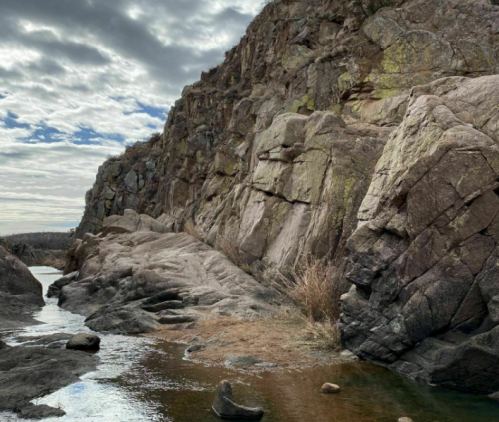Rocky cliffside with a calm water body in the foreground, under a cloudy sky.
