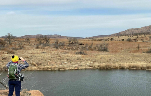 A person in a yellow beanie takes a photo of a scenic landscape with a river and distant hills.