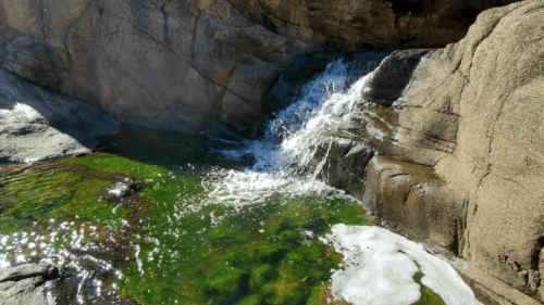 A small waterfall cascades over rocks into a clear, green pool surrounded by smooth stone surfaces.
