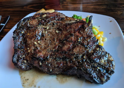 A large, grilled ribeye steak on a white plate, garnished with herbs and accompanied by mixed vegetables.