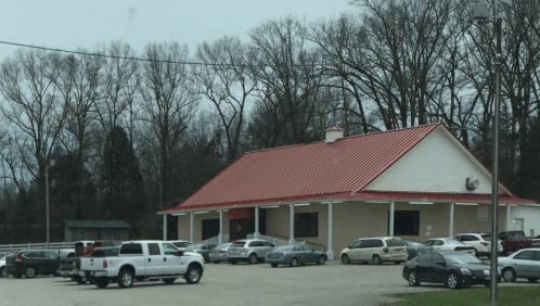 A building with a red roof surrounded by parked cars and bare trees in the background on a cloudy day.