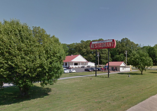 A roadside restaurant with a red sign, surrounded by trees and parked cars, on a sunny day.