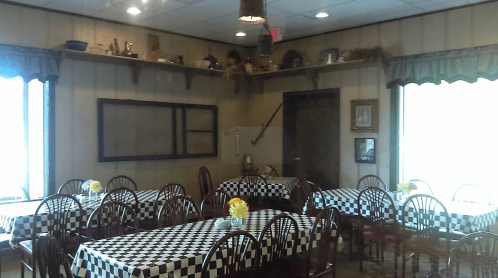 Cozy dining area with checkered tablecloths, wooden chairs, and decorative shelves, featuring flowers on each table.